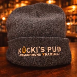 Dark grey knit beanie with embroidered “Kücki’s Pub – Swakopmund, Namibia” logo on a wooden bar counter in a warm pub setting.