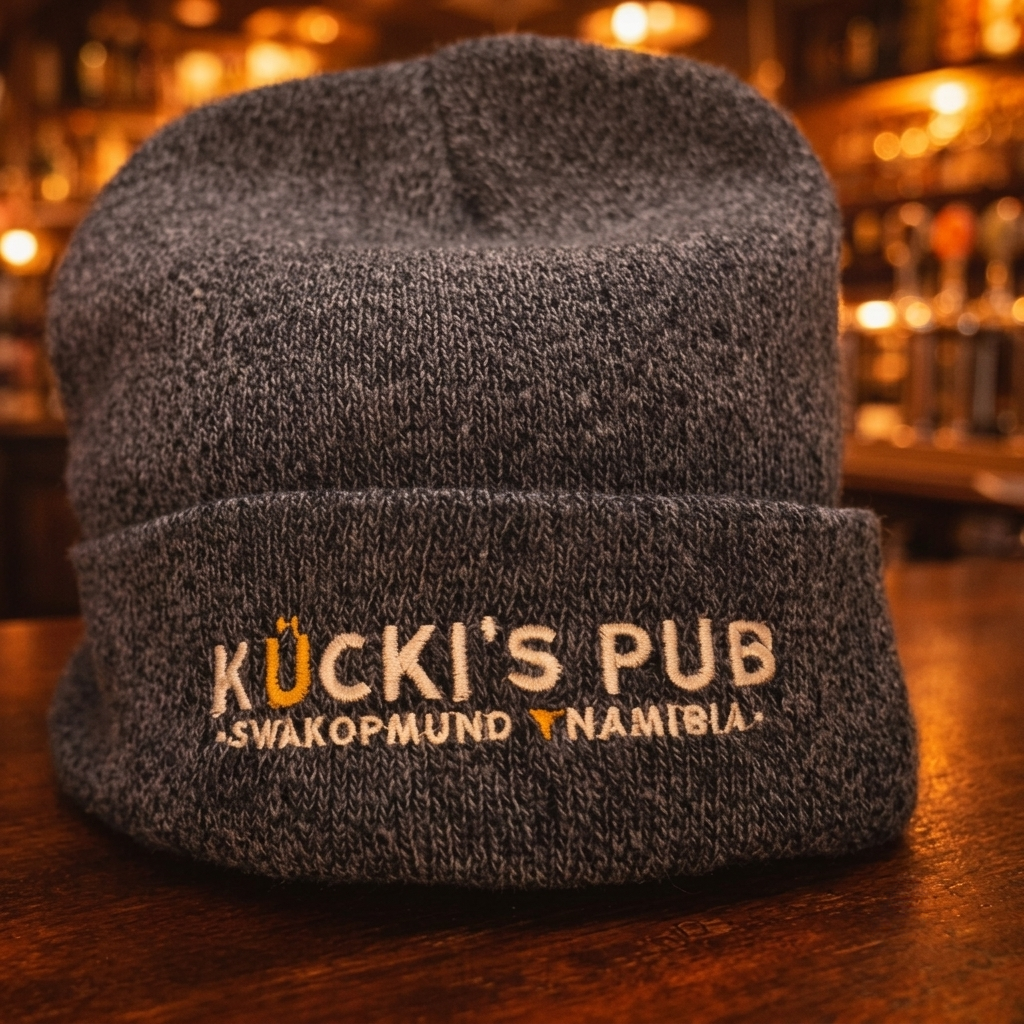Dark grey knit beanie with embroidered “Kücki’s Pub – Swakopmund, Namibia” logo on a wooden bar counter in a warm pub setting.