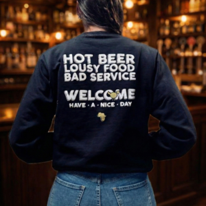 Rear view of a woman with a French braid in blue jeans and a black sweatshirt against a blurred bar background with warm hanging lights. The sweatshirt displays humorous text: 'HOT BEER LOUSY FOOD BAD SERVICE WELCOME HAVE • A • NICE • DAY'.
