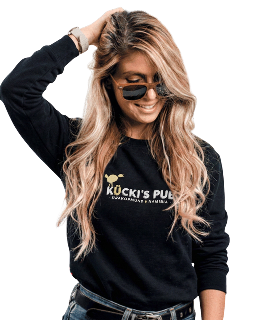 A blonde woman in sunglasses and a black "Kücki's Pub" sweatshirt, smiling with her hand in her hair, against a white and pink geometric background.