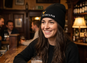 A smiling young woman with long, wavy brown hair sitting at a rustic wooden bar, wearing a black knit beanie featuring the "Kücki’s Pub" logo and a yellow bird emblem. She is holding a glass of beer, with a warm, blurred pub background including bottles and a vintage lamp.