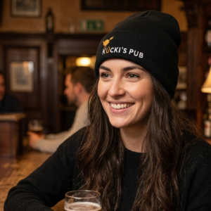 A smiling young woman with long, wavy brown hair sitting at a rustic wooden bar, wearing a black knit beanie featuring the "Kücki’s Pub" logo and a yellow bird emblem. She is holding a glass of beer, with a warm, blurred pub background including bottles and a vintage lamp.