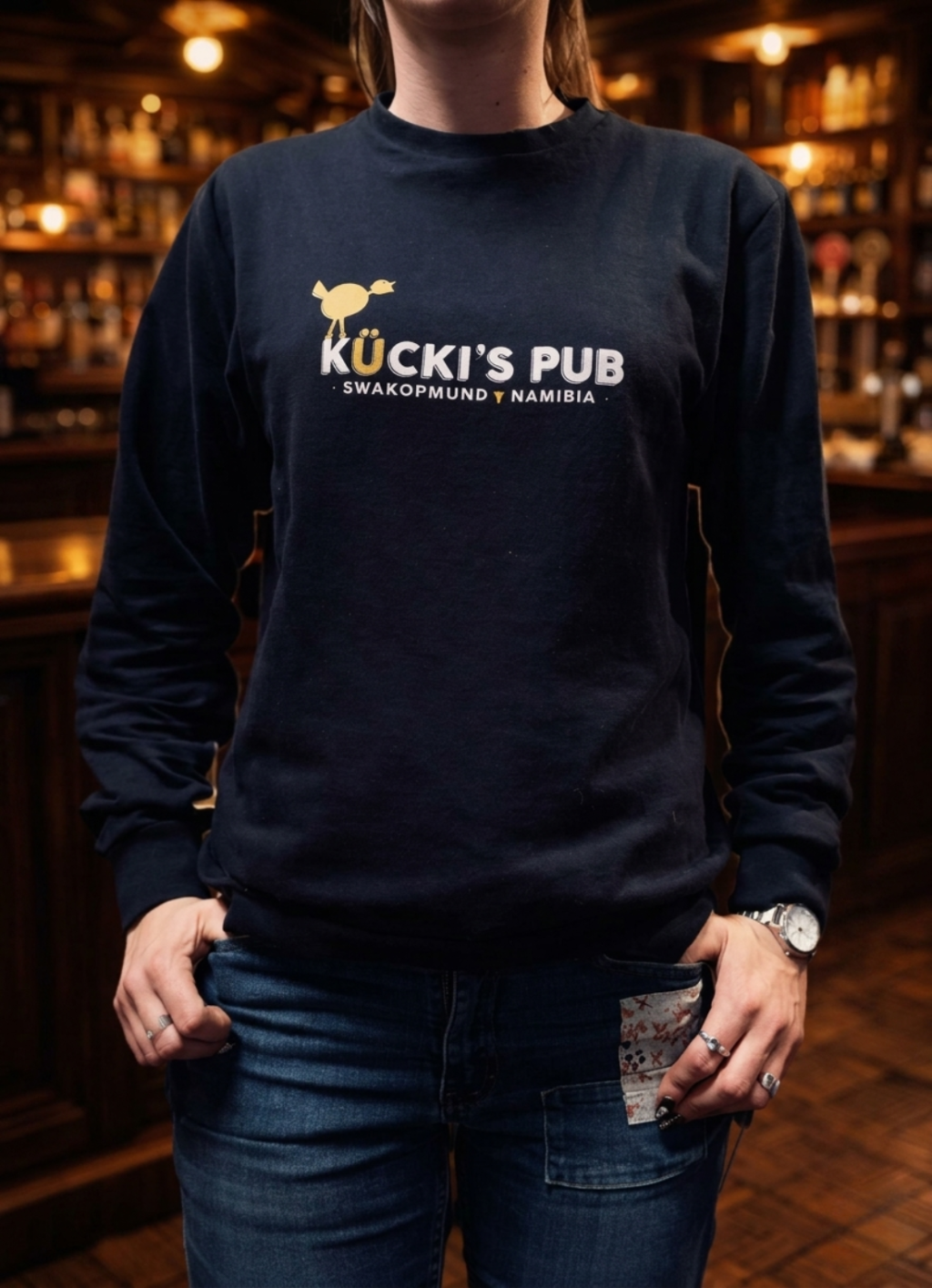 Woman wearing a black Kücki’s Pub sweatshirt with logo, standing in a warm, traditional pub setting.